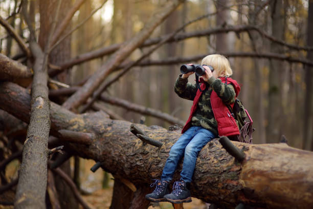 Little boy scout with binoculars during hiking in autumn forest. Child is sitting on large fallen tree and looking through a binoculars. Concepts of adventure, scouting and hiking tourism for kids.